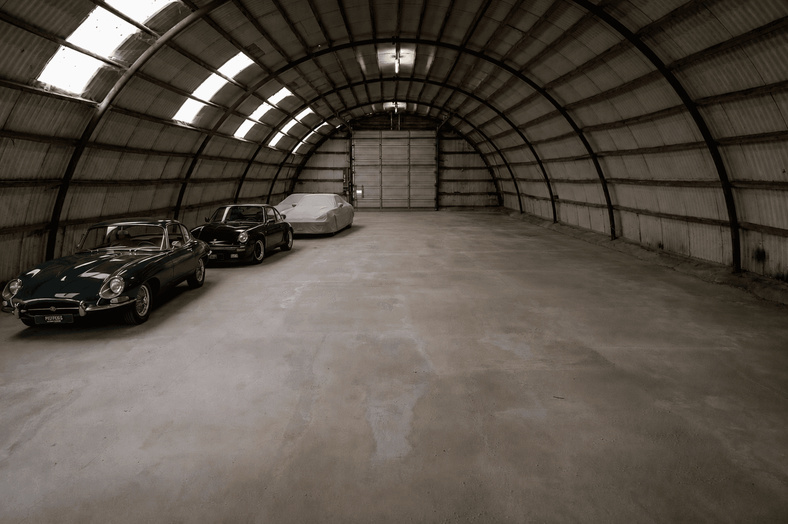 A green Jaguar and black Porsche parked in a spacious, arched metal storage hangar.